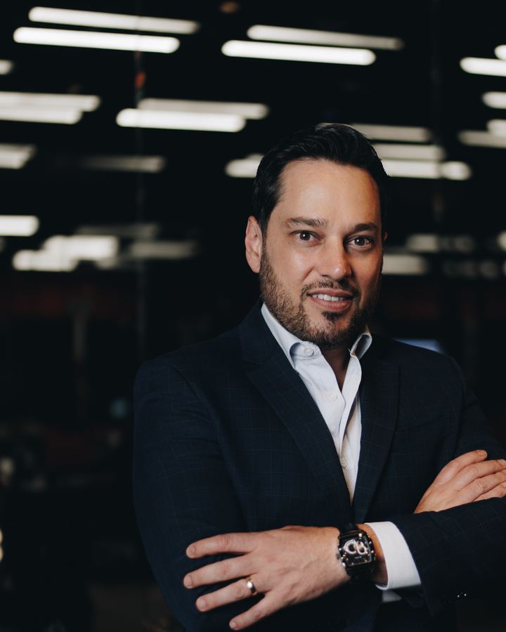 Lucien Newton de terno escuro, braços cruzados, em um ambiente corporativo